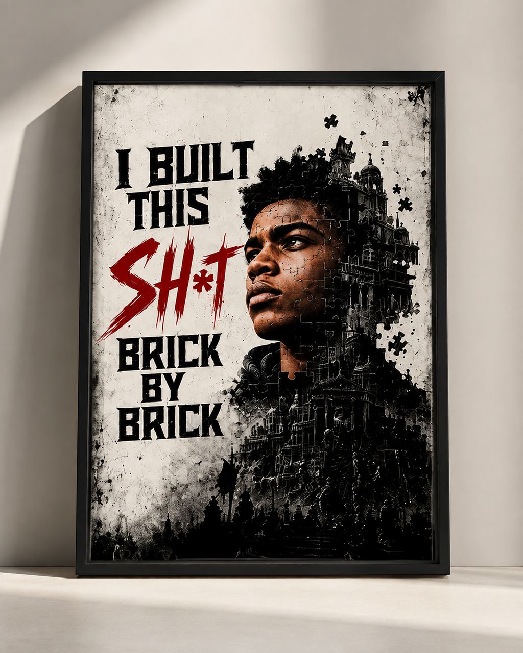 A framed black-and-white graphic print leaning against a light wall. The artwork features the text 'I BUILT THIS SH*T BRICK BY BRICK' in black and red, with a stylized portrait of a man integrated into a brick-and-building collage.