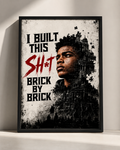 A framed black-and-white graphic print leaning against a light wall. The artwork features the text 'I BUILT THIS SH*T BRICK BY BRICK' in black and red, with a stylized portrait of a man integrated into a brick-and-building collage.