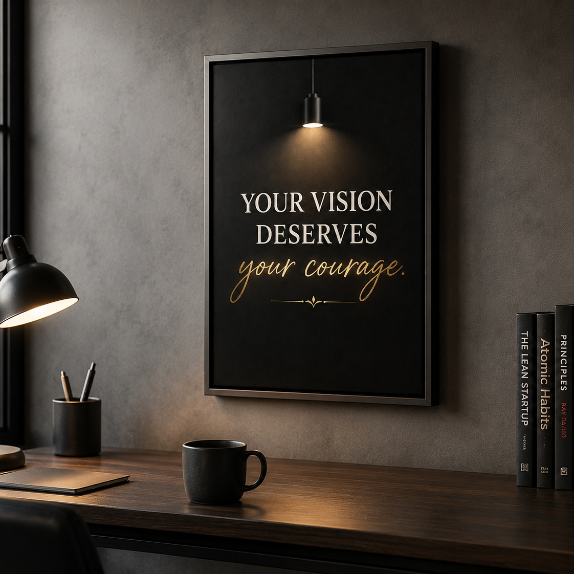 A framed motivational wall art print hanging on a gray wall above a wooden desk. The print has a black background with the text 'YOUR VISION DESERVES your courage.