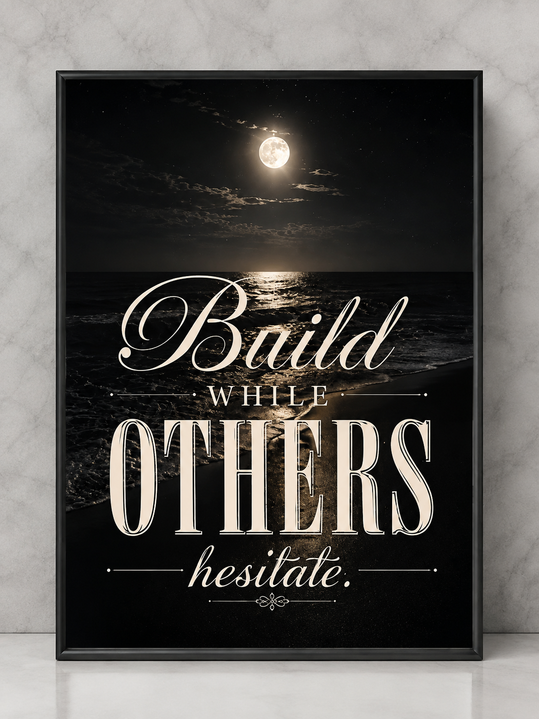 A framed motivational art print featuring a night ocean scene with a full moon and the quote 'Build WHILE OTHERS hesitate.' in cream lettering, shown leaning against a light gray marble wall.